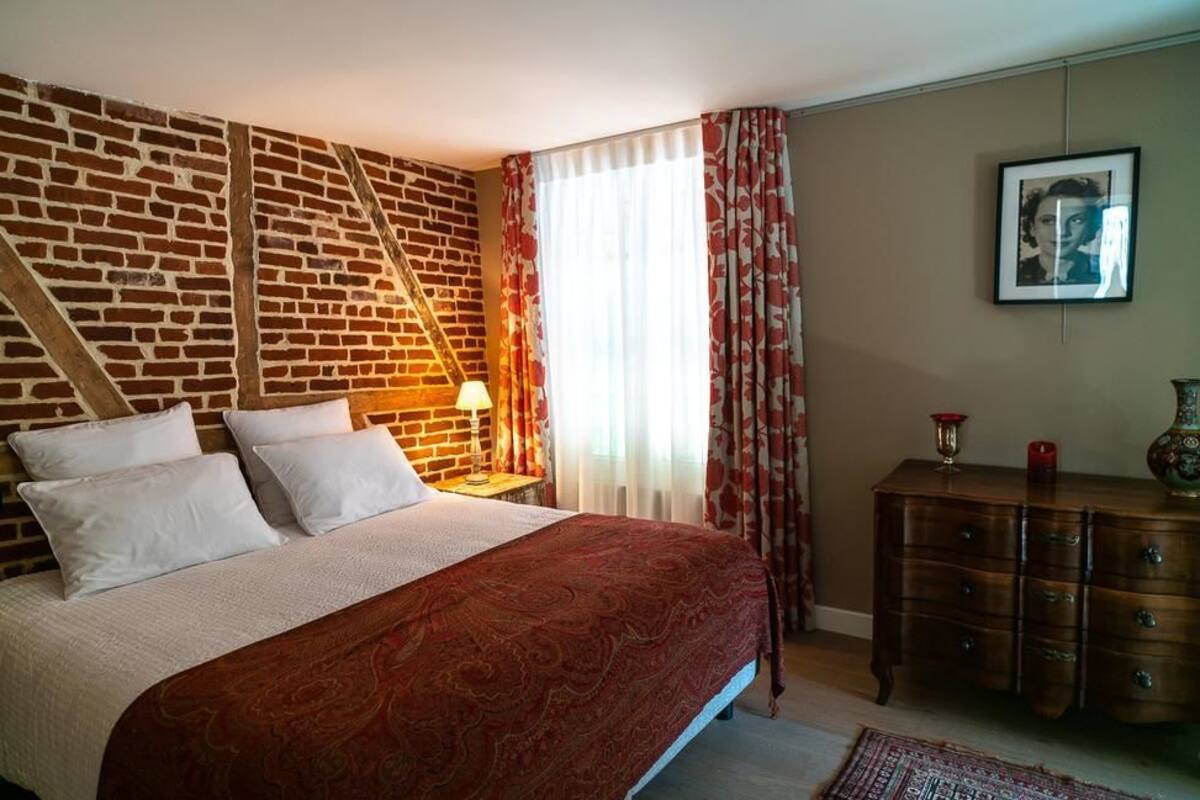 Un dormitorio con pared de ladrillo, cama con ropa de cama blanca y roja, mesita de noche con lámpara, cómoda y un cuadro enmarcado en la pared.