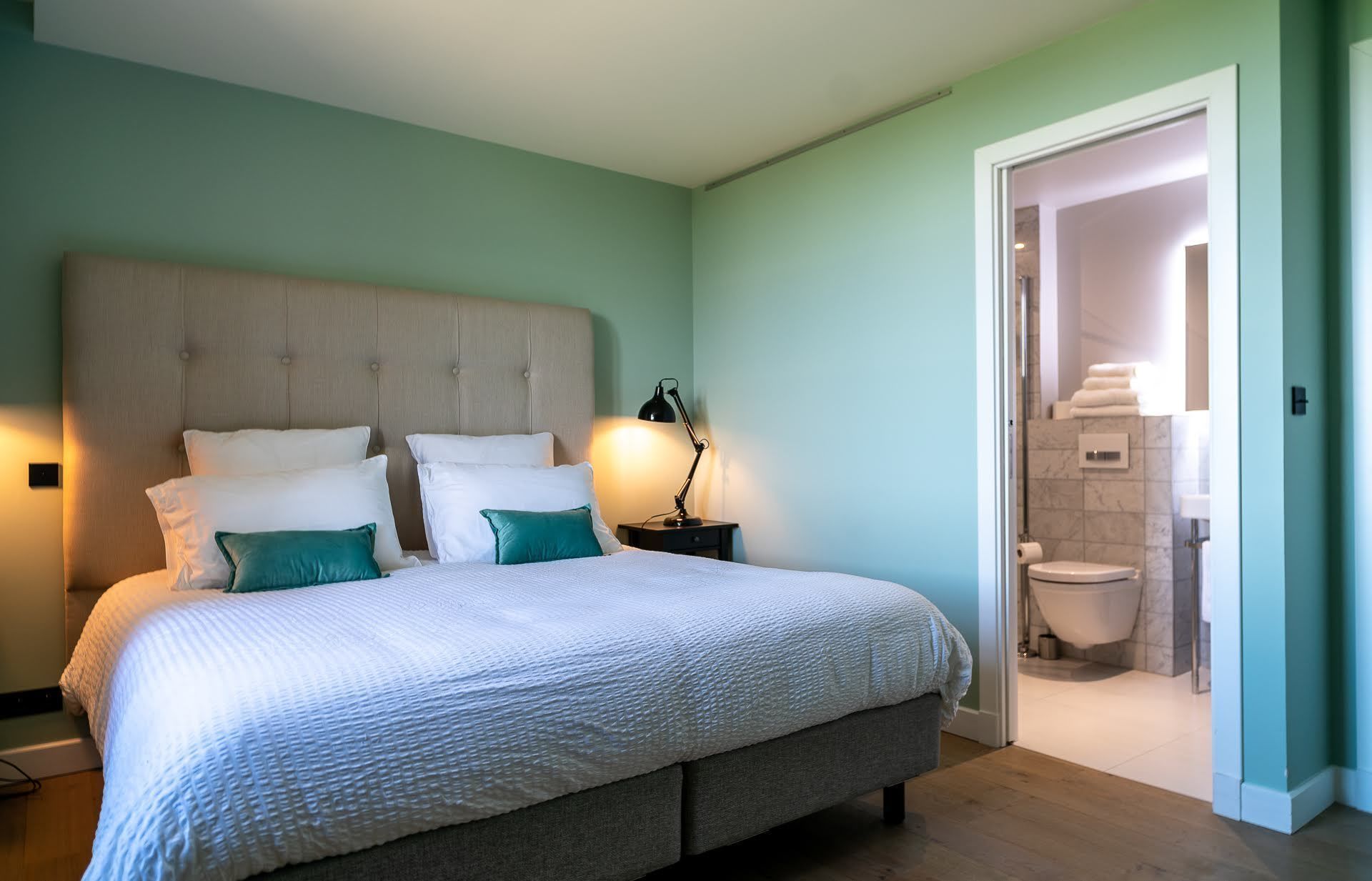 Ein Schlafzimmer mit einem großen Bett, einem Nachttisch und einer Lampe. Ein Badezimmer ist durch eine offene Tür sichtbar.