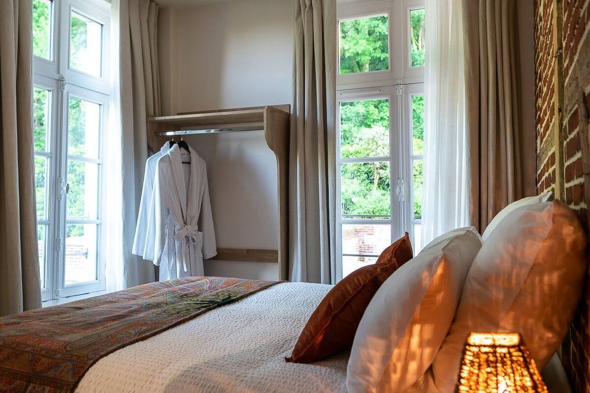 Un dormitorio con una cama grande, una mesita de noche con lámpara, una ventana con cortinas y una bata colgada en un perchero.