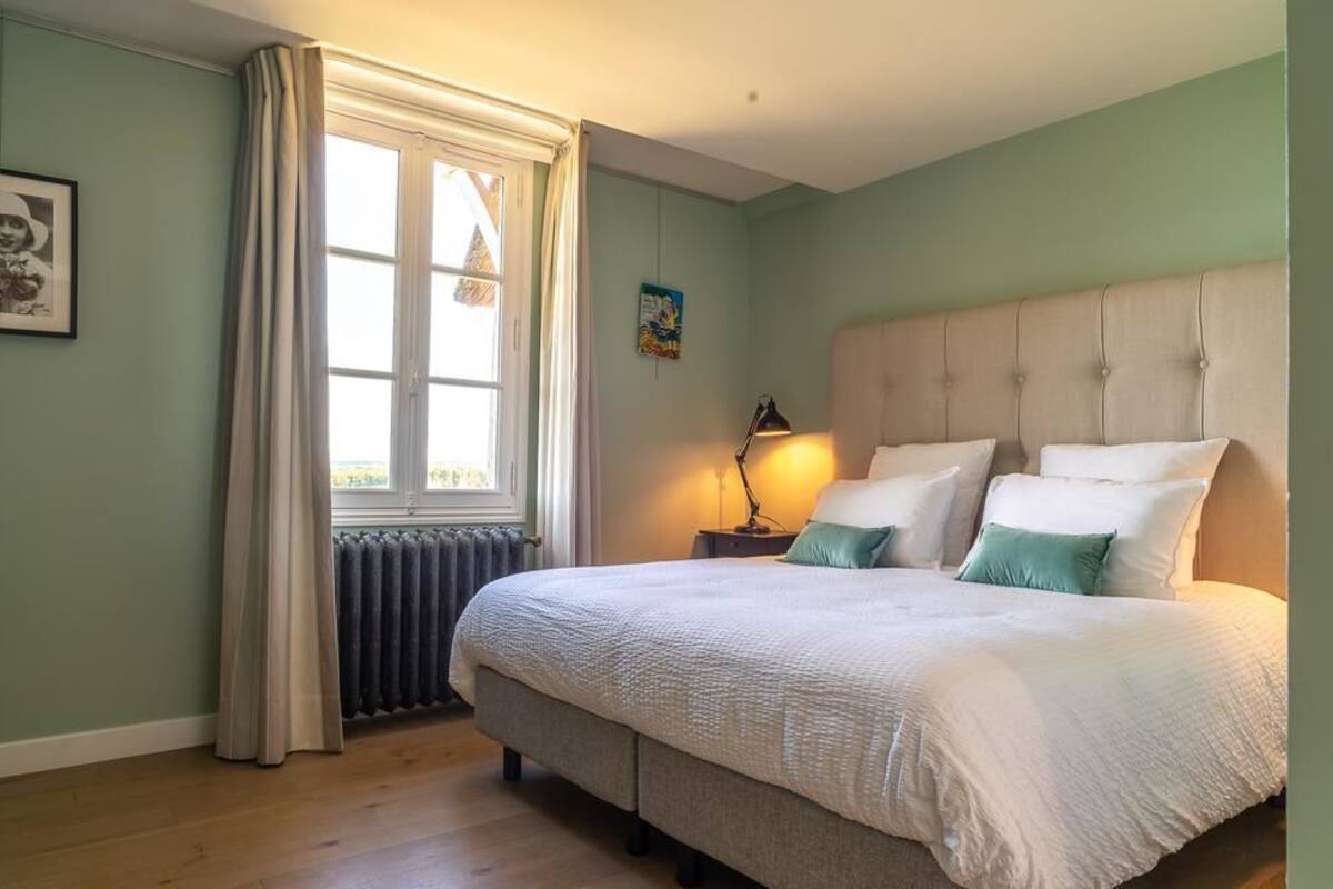 Un dormitorio con una cama grande, una ventana con cortinas, un radiador y una mesita de noche con lámpara.
