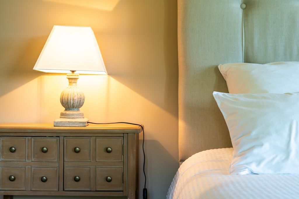 Een keurig opgemaakt bed met witte lakens en kussens, een bedlampje op een houten nachtkastje met lades, en een effen muur met een gordijn.