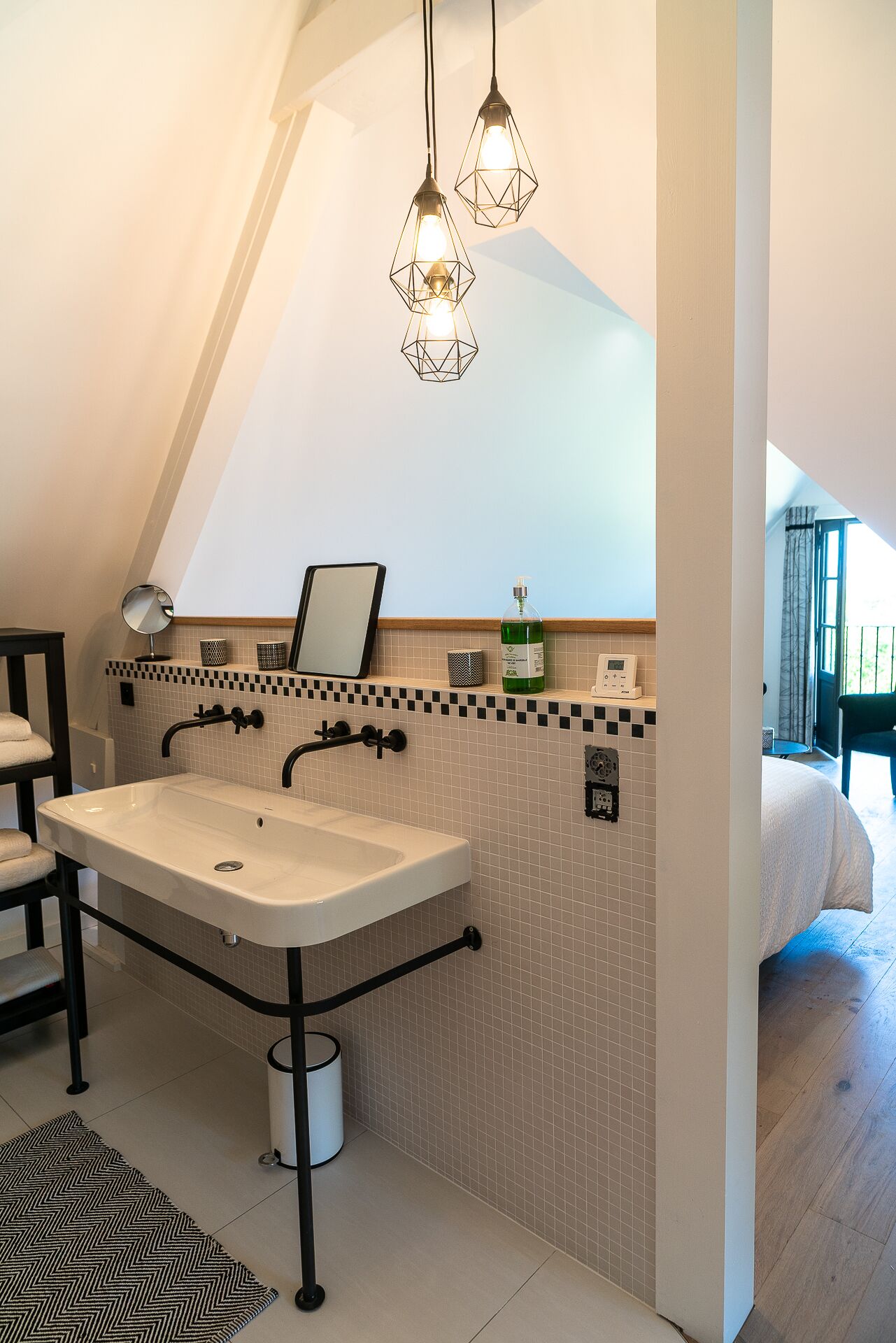 A modern bathroom with a large sink, two faucets, and a mirror. There are hanging lights, a small shelf with toiletries, and a tiled backsplash.