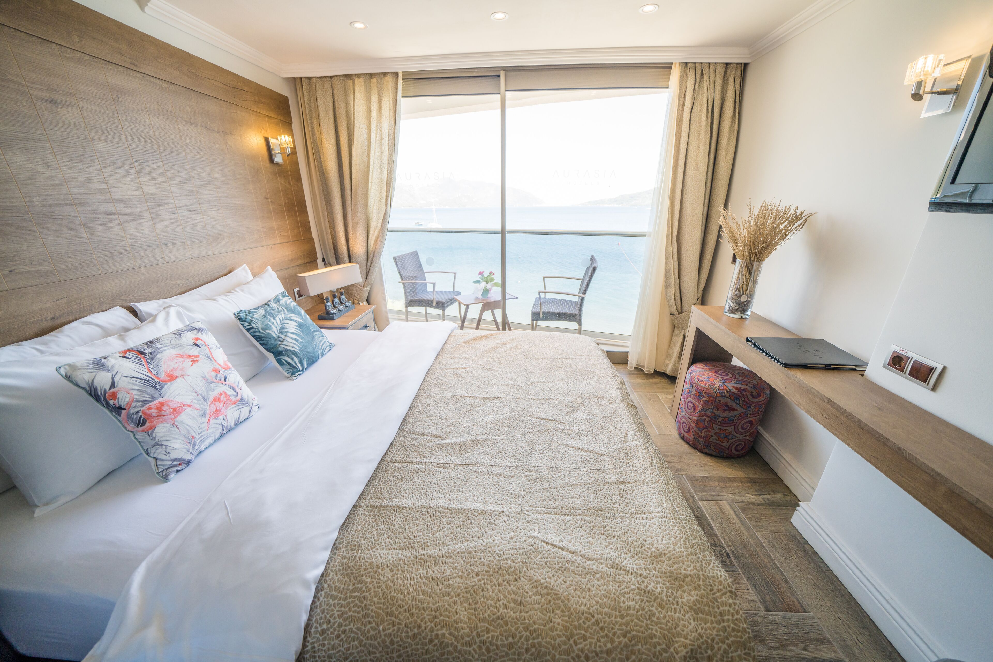 Habitación de hotel con una cama grande, un escritorio con una computadora portátil, una silla y vista al mar a través de un ventanal amplio.