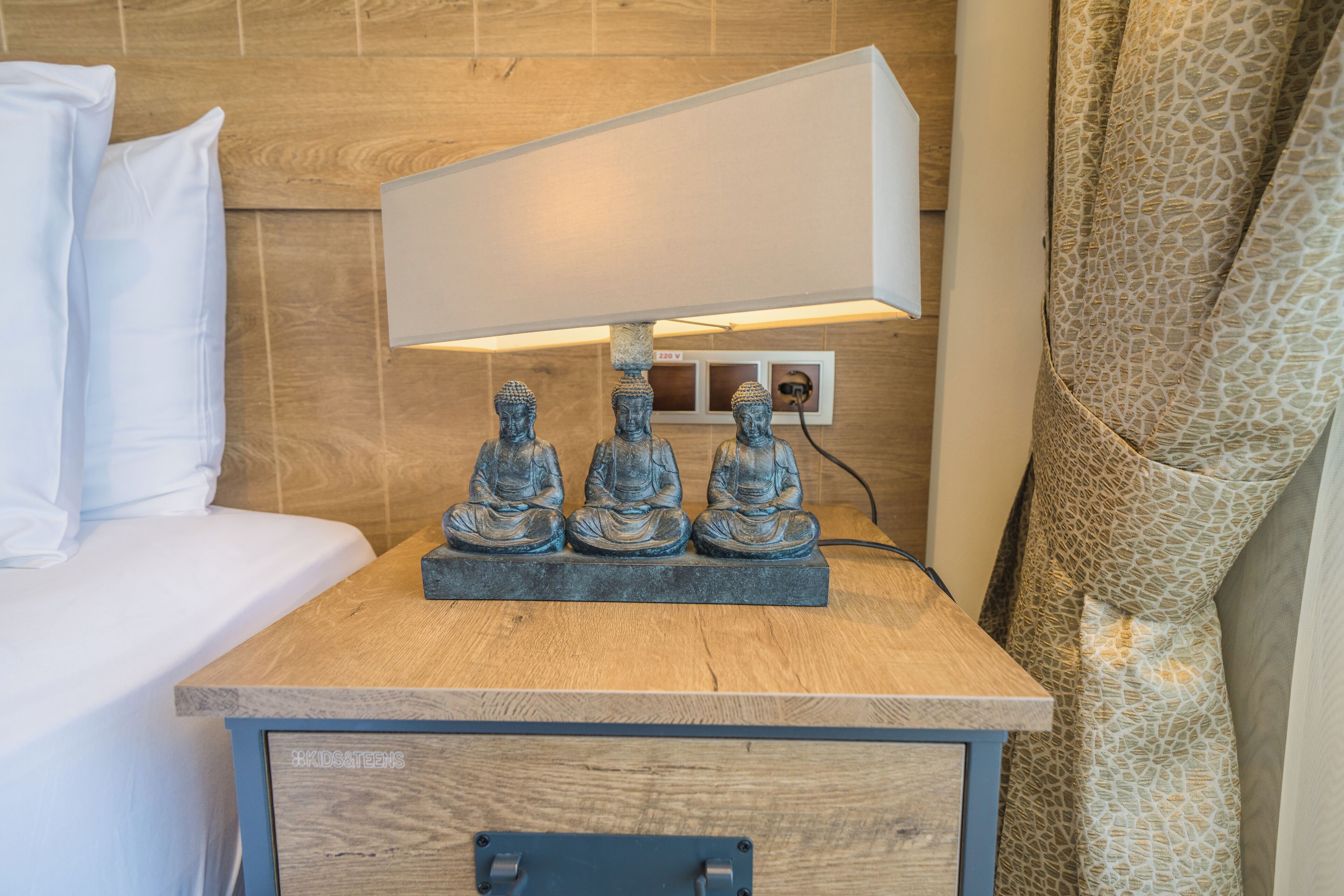Ein Nachttisch aus Holz mit einer Lampe, drei Buddha-Statuen und einer kleinen Schublade.
