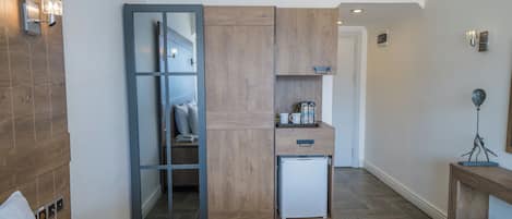 A compact hotel room with a wooden wardrobe, a small kitchenette, and a bedside table with a phone.