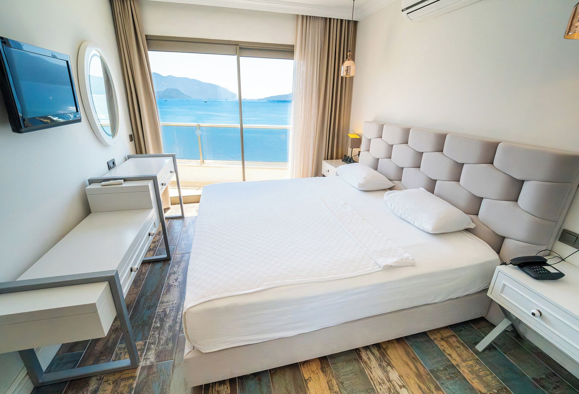 A hotel room with a large bed, a desk, a TV, and a view of the sea.