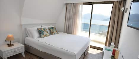 A hotel room with a large bed, a nightstand with a lamp, a TV, and a view of the sea.