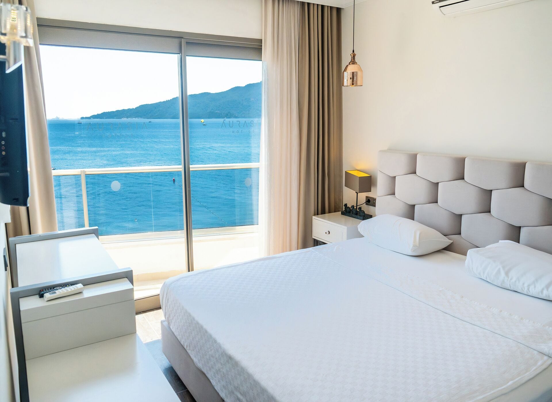 A hotel room with a bed, a bedside table, a lamp, a TV, and a view of the sea.