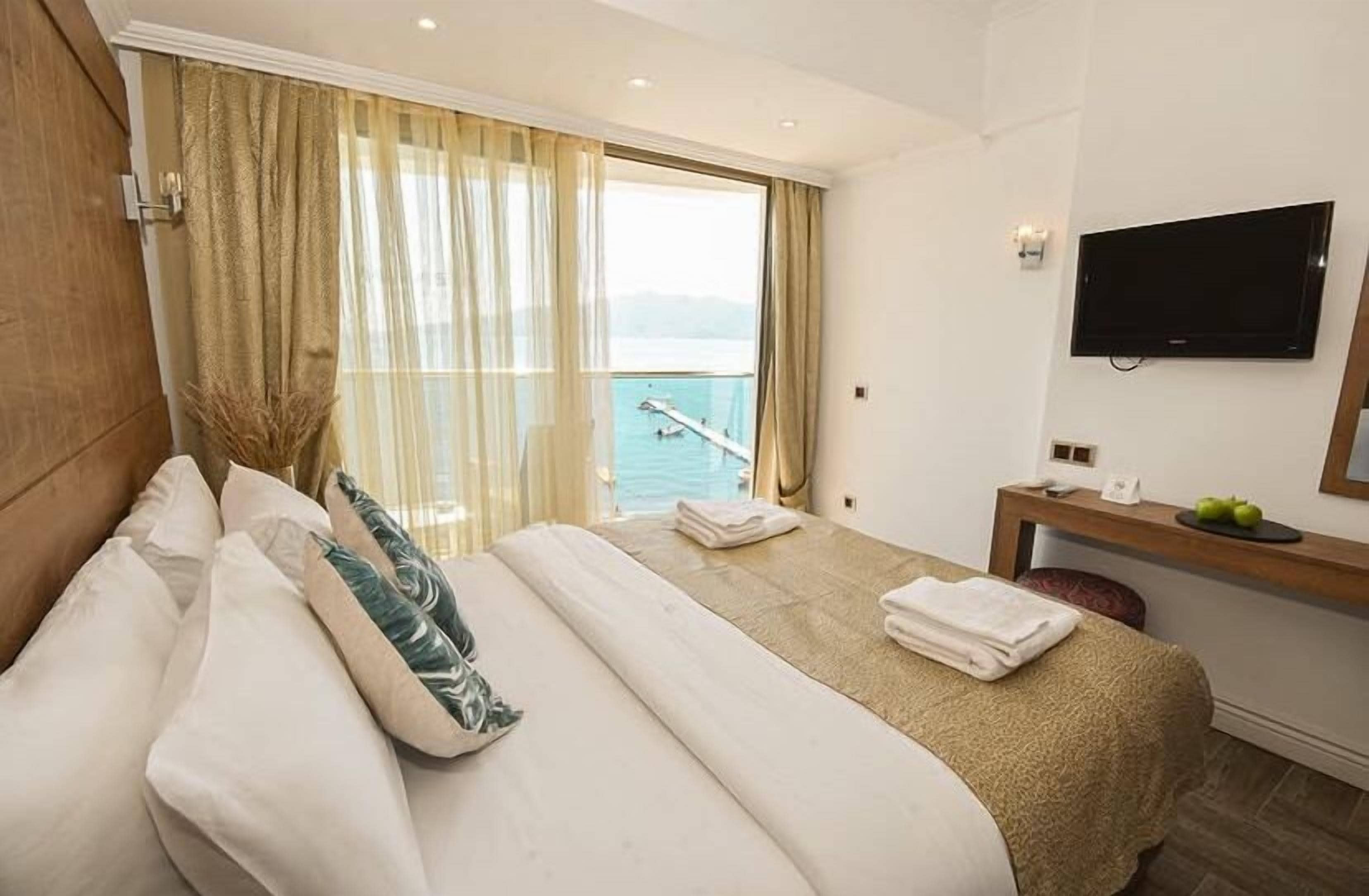 A hotel room with a bed, a desk, a TV, and a view of the sea.