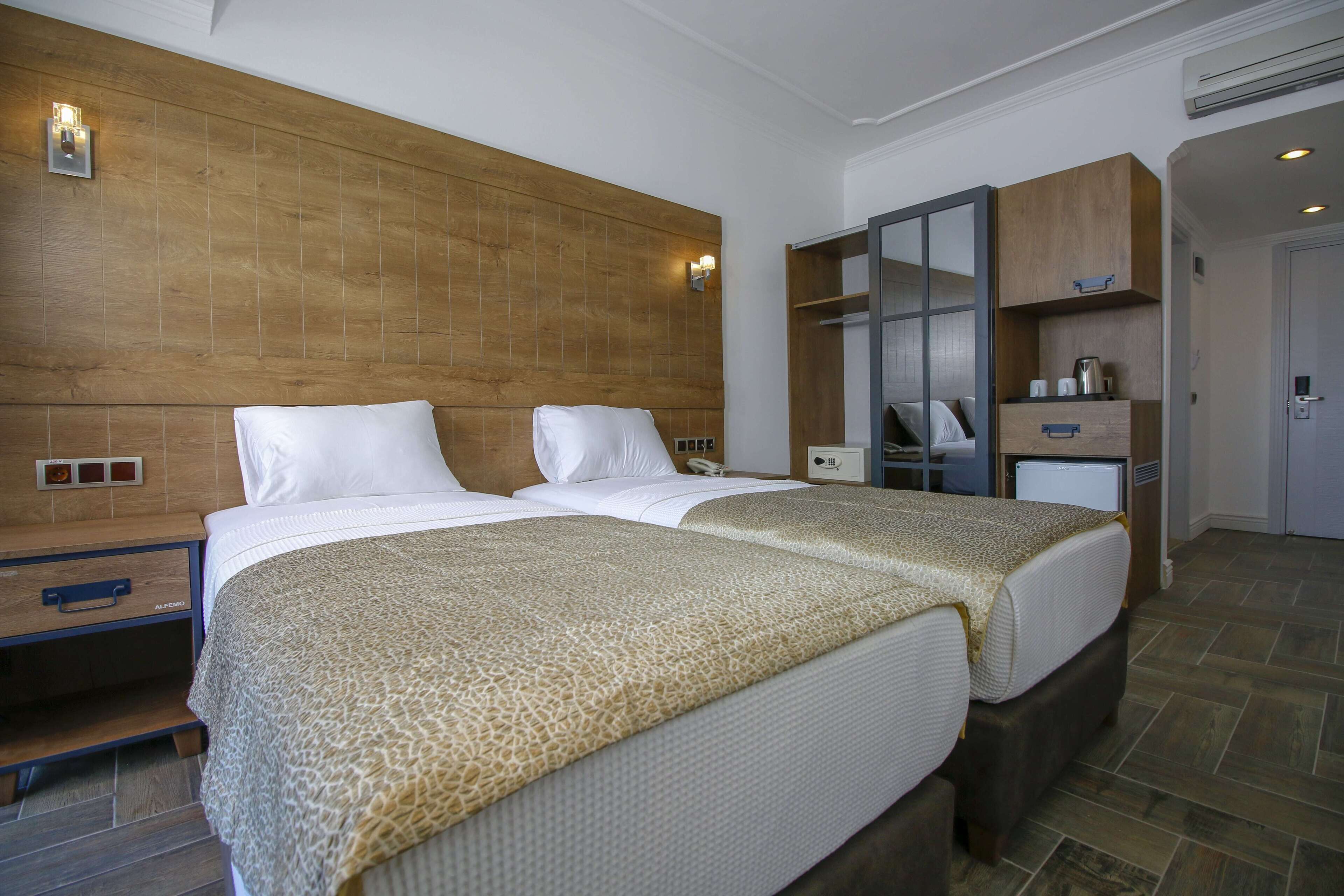 A hotel room with two beds, a wooden headboard, a nightstand, a wardrobe, and a small desk with a lamp.
