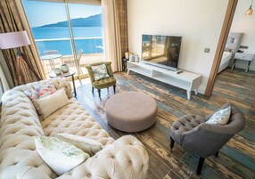 A living room with a large sofa, ottoman, armchair, TV, and a view of the sea.