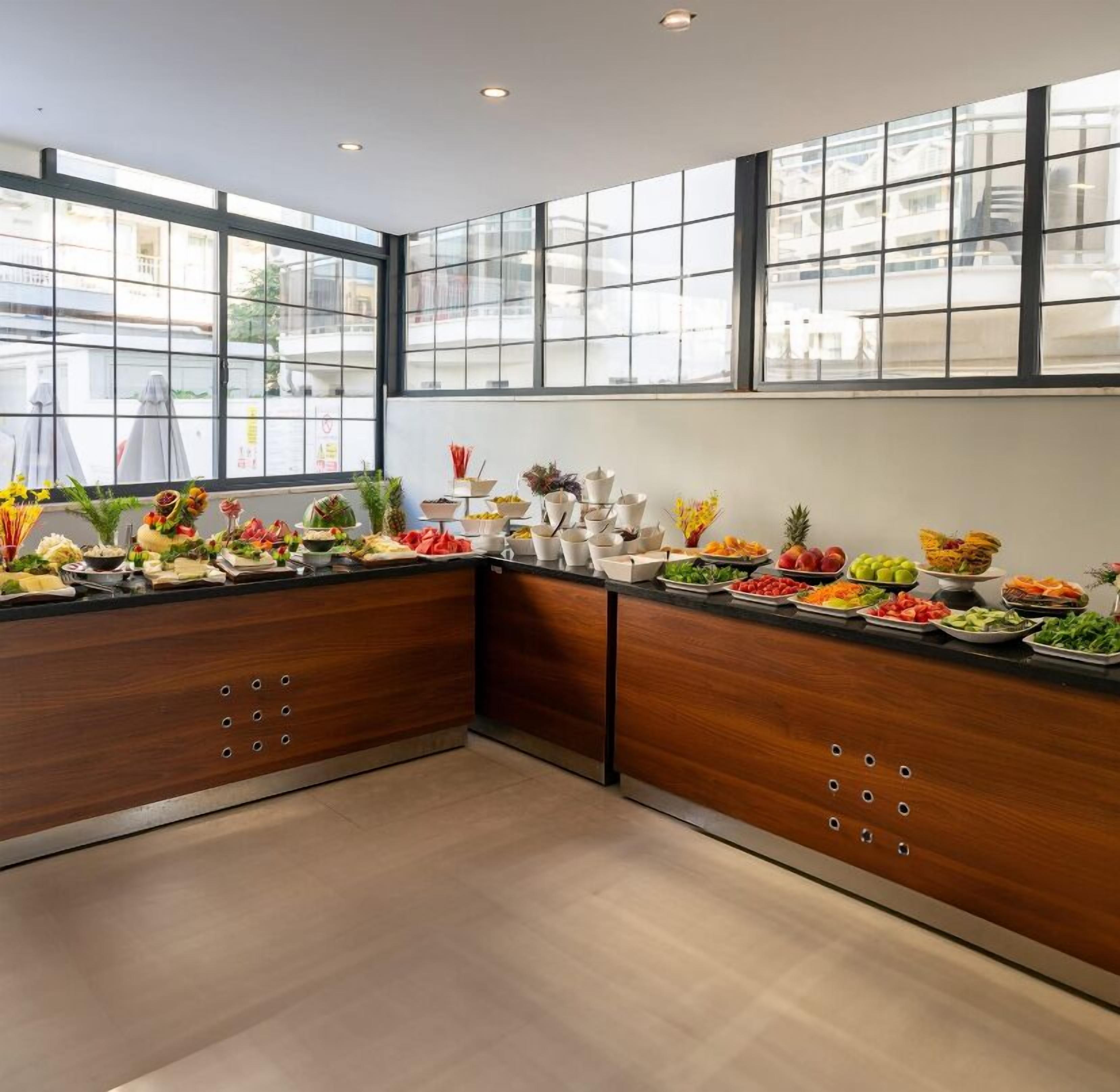 A modern kitchen with wooden cabinets and a glass partition, featuring a buffet with various fruits and vegetables.