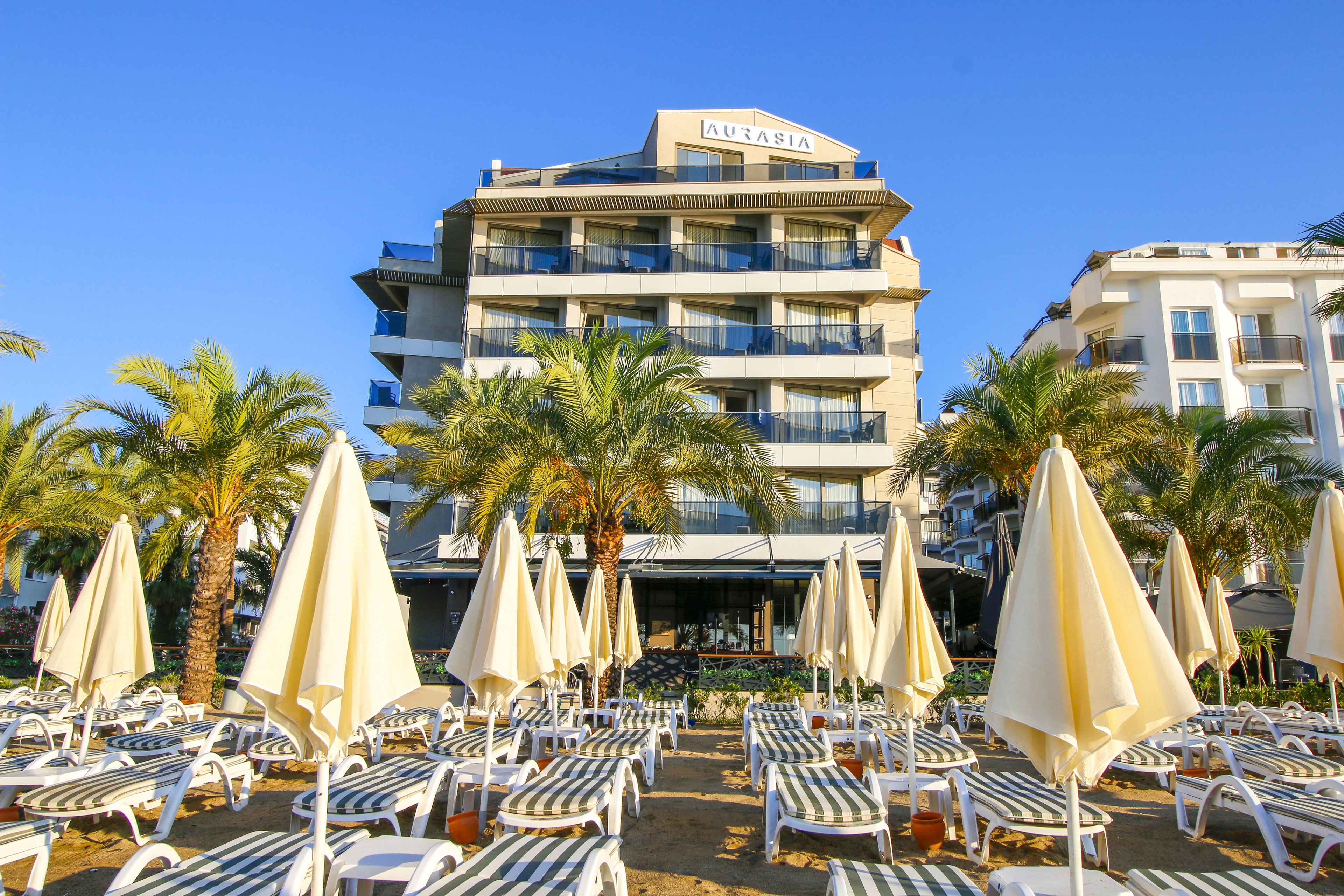 Un hôtel appelé « AURASIA », doté de nombreux balcons, de chaises longues et de palmiers en premier plan.