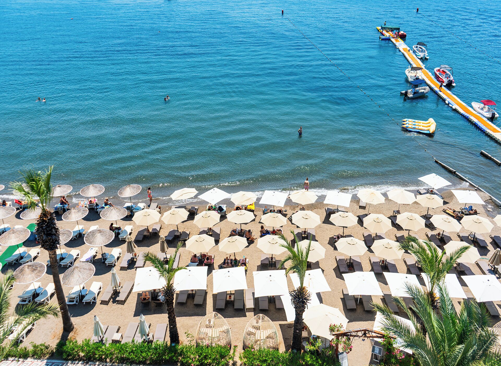 Una playa con filas de sillas de sol y sombrillas, un muelle con barcos, y una vista despejada del mar.