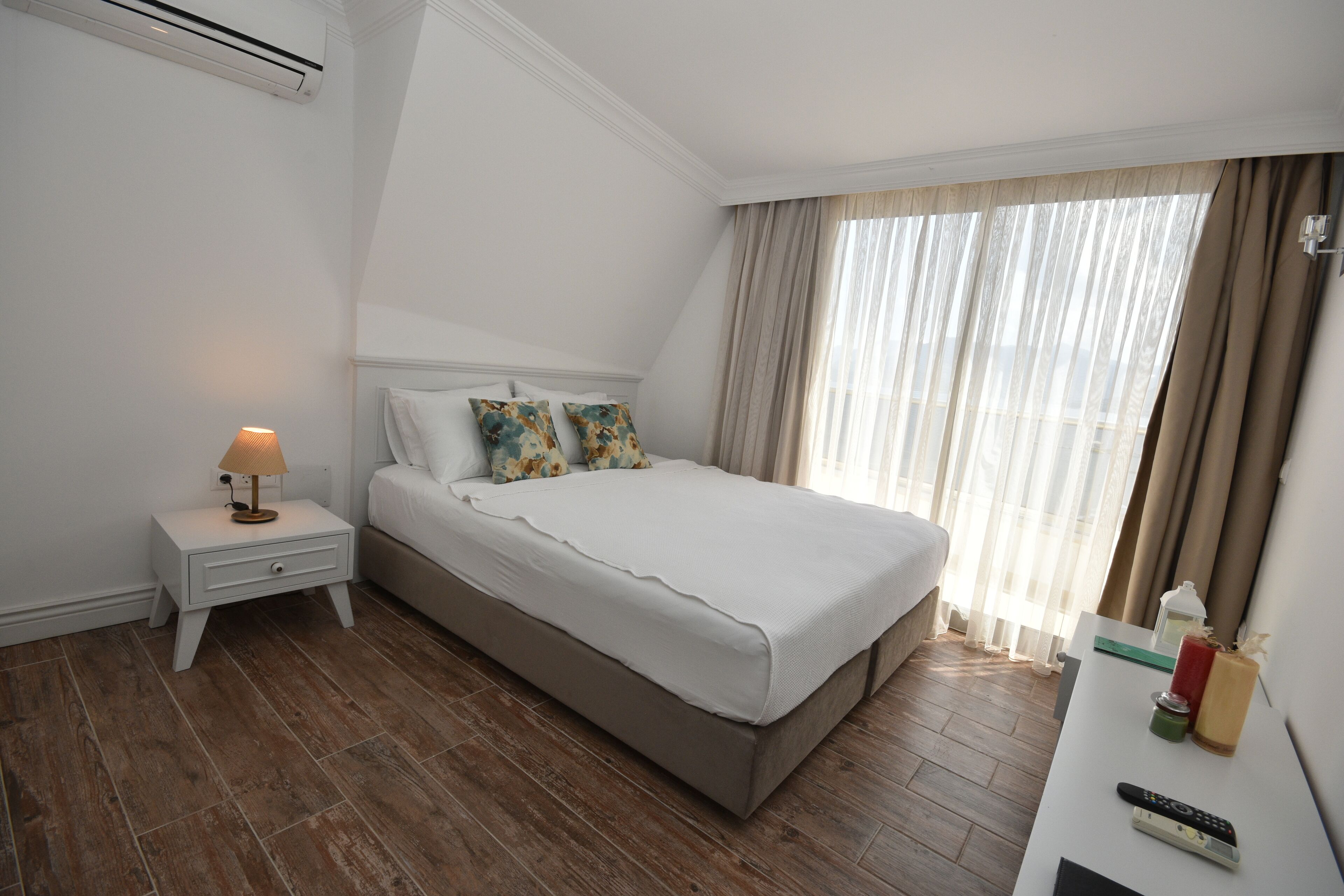 A hotel room with a bed, a nightstand, a lamp, a remote control, and a window with curtains.