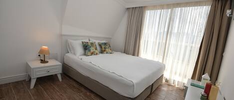 A hotel room with a bed, a nightstand, a lamp, a remote control, and a window with curtains.