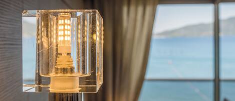Un candelabro moderno con estructura de vidrio transparente, una fuente de luz visible y una vista a un balcón con barandilla y un paisaje lejano.