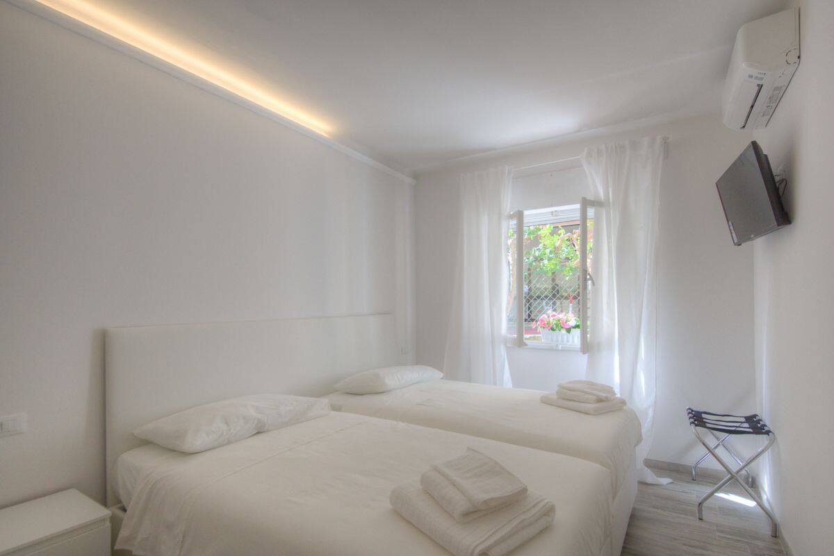 Ein Hotelzimmer mit zwei Betten, einem Fenster mit Vorhängen, einem an der Wand befestigten Fernseher und einem kleinen Tisch mit Stuhl.