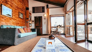 A cozy living room with a wooden wall, a blue sofa, a coffee table with a GT HOUSE sign, and a large window with a view. - Casa da Rampa By Gt House (Galamares)