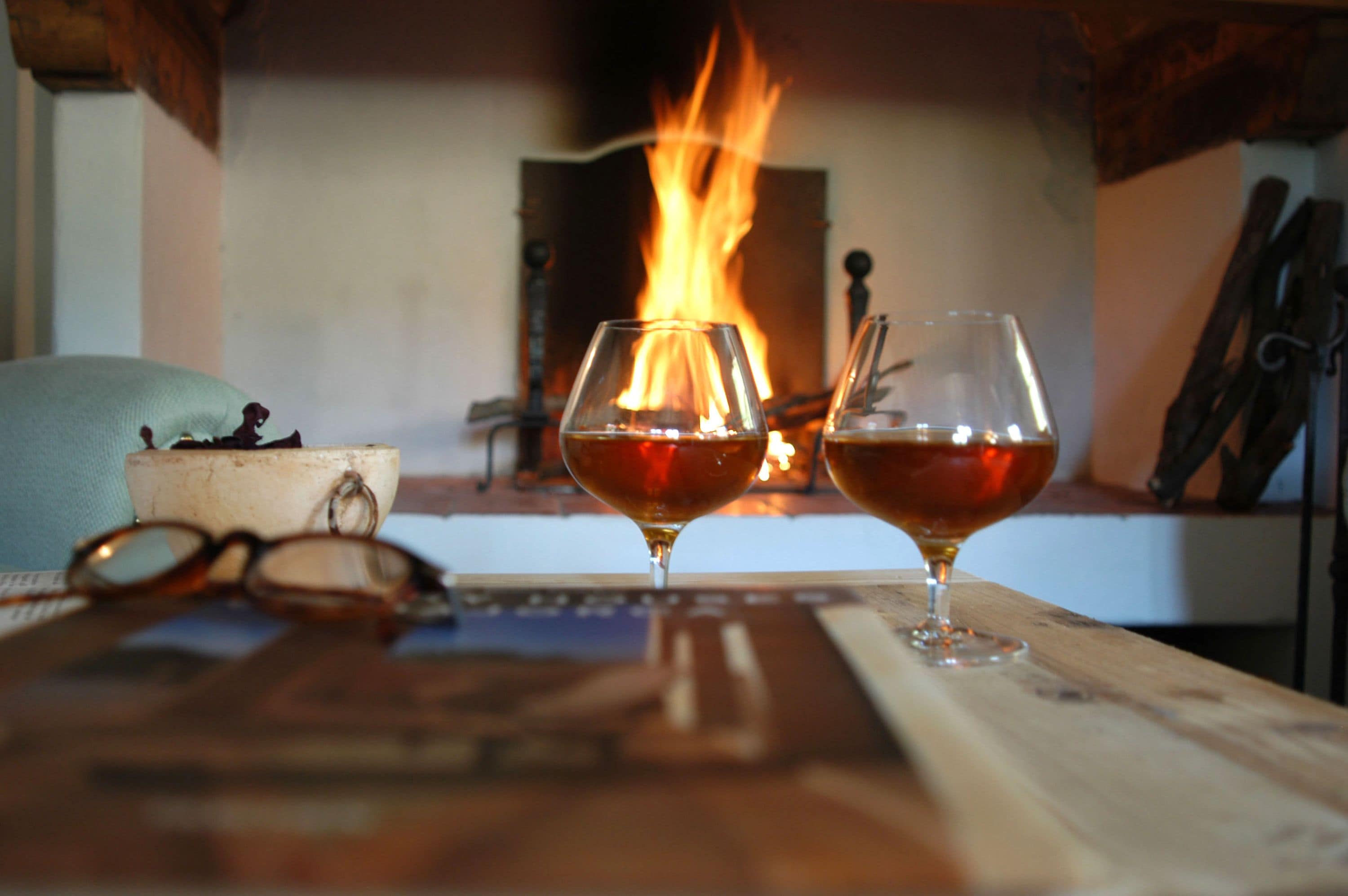 Deux verres contenant un liquide ambré sont posés sur une table en bois, avec une cheminée en arrière-plan.