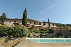 A pool area with lounge chairs and umbrellas, a stone building with a tiled roof, and a clear blue sky. - Il Defizio (Gambassi Terme)