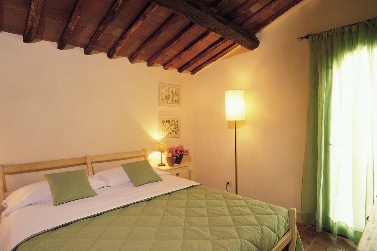 A neatly made bed with a green quilt, a bedside lamp, and a small table with a flower vase.