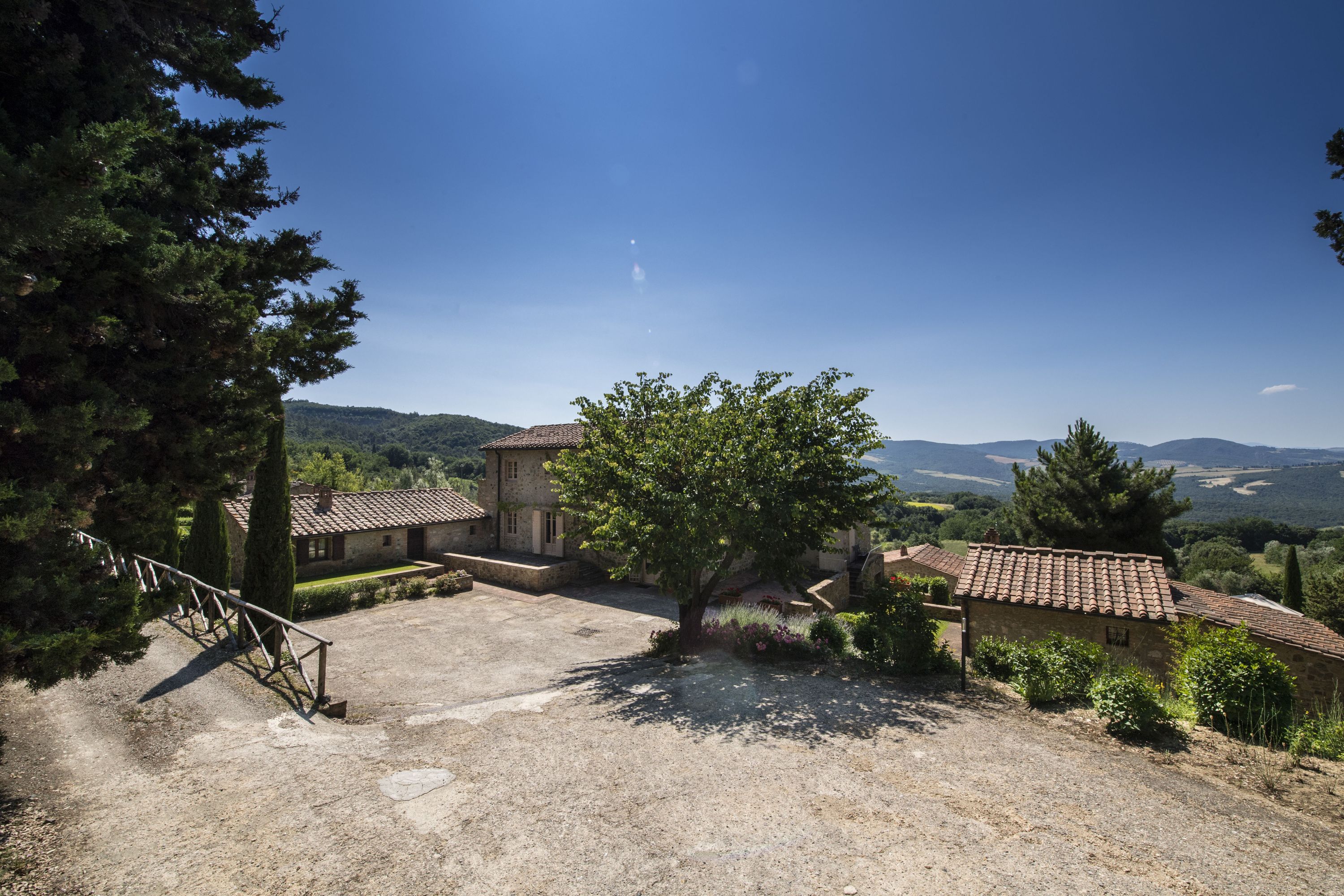 A rustic stone building with a tiled roof, surrounded by a gravel courtyard and a large tree.
