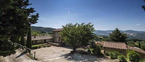 A rustic stone building with a tiled roof, surrounded by a gravel courtyard and a large tree.