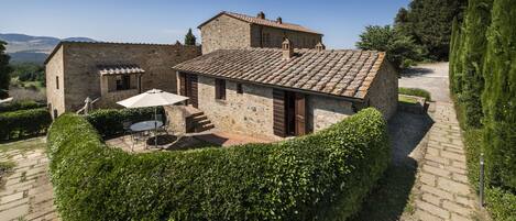 A stone building with a tiled roof, a patio with outdoor furniture, and a neatly trimmed hedge.
