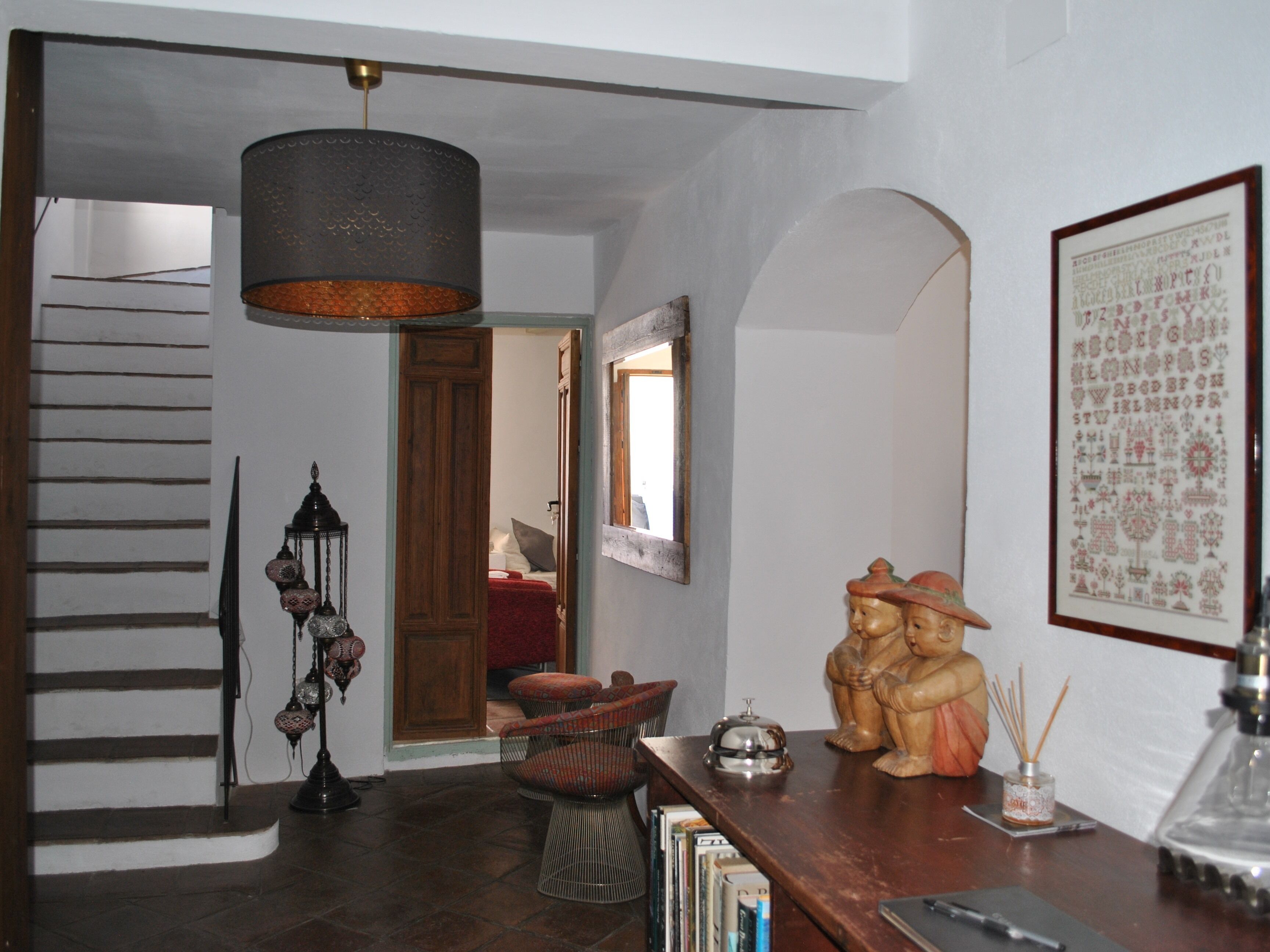 Un escalier avec une rampe en bois, une lampe suspendue de couleur sombre, une lanterne décorative, une petite table surmontée d’une statue, une œuvre d’art textile encadrée et une bibliothèque.