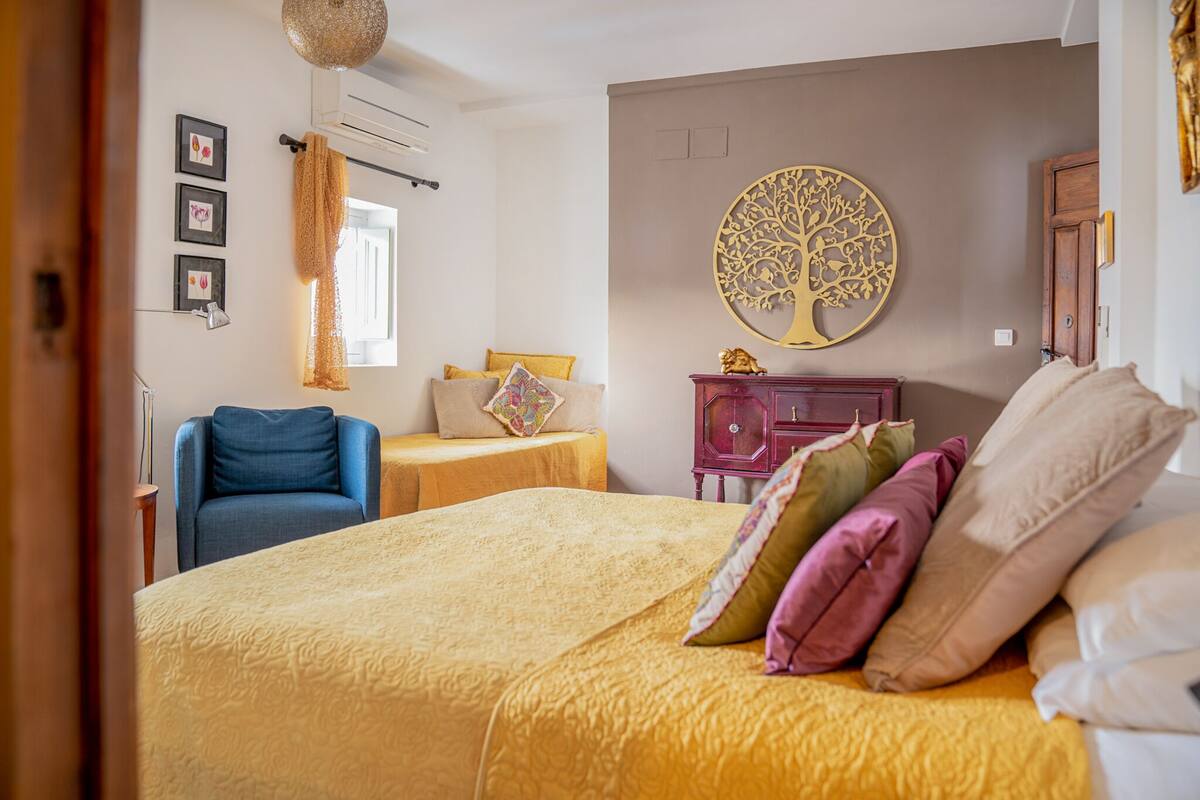 A bedroom with a large bed, a chair, a small table, a dresser, and a wall decoration.