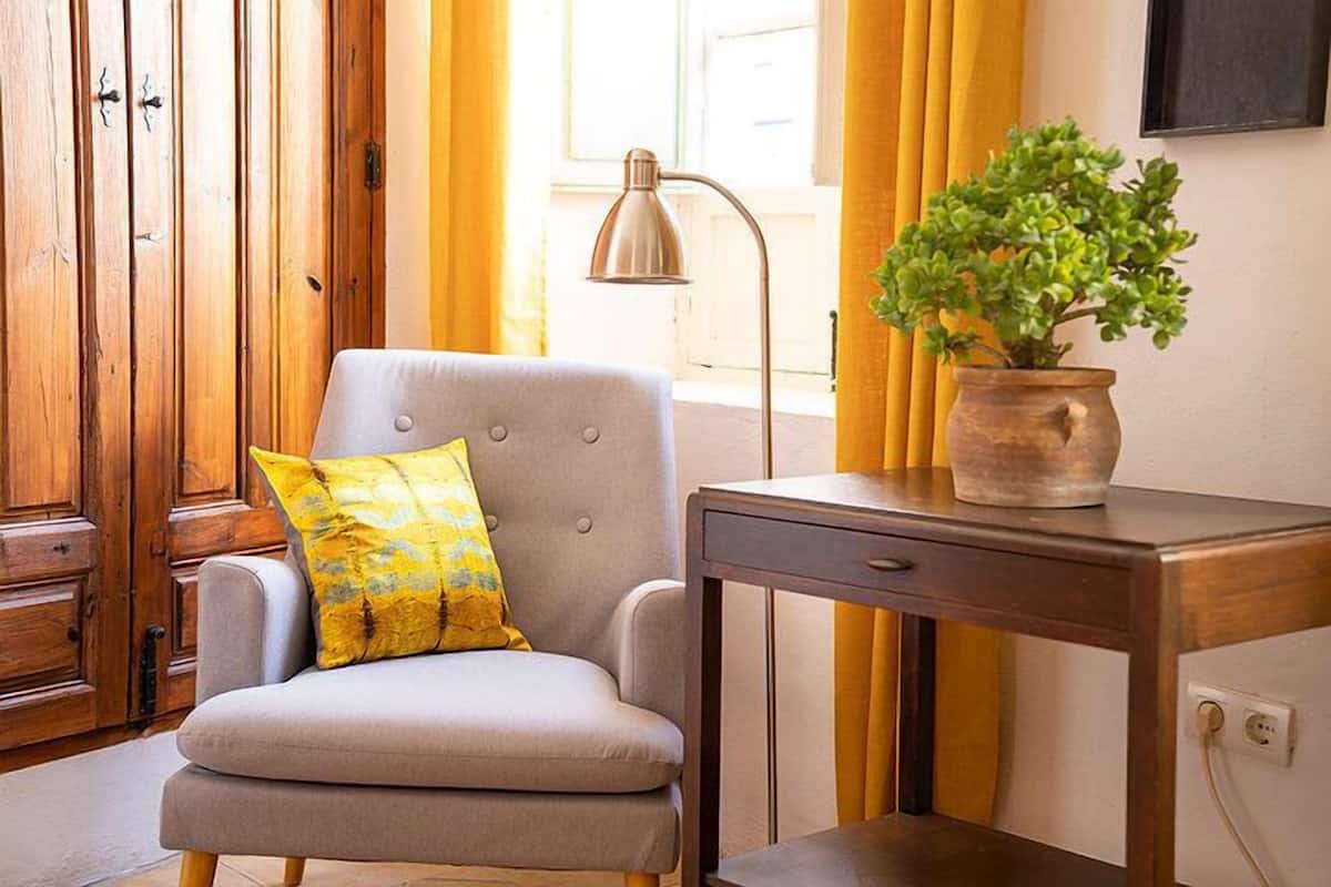 A room with a grey armchair, a wooden cabinet, a desk with a potted plant, and a lamp.