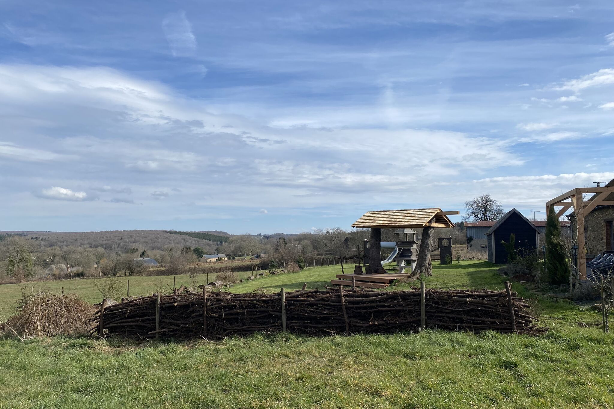 Un paysage rural avec une structure en bois, un tas de branches et une vue lointaine sur des collines.