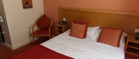 A neatly made bed with a red quilt, a wooden headboard, and two pillows. There is a chair with a red cushion and a bedside table with a phone and a lamp.