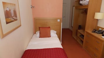 A single bed with a wooden headboard, a red bedspread, and a red pillow. There is a wooden wardrobe with a mirror and a lamp on top.