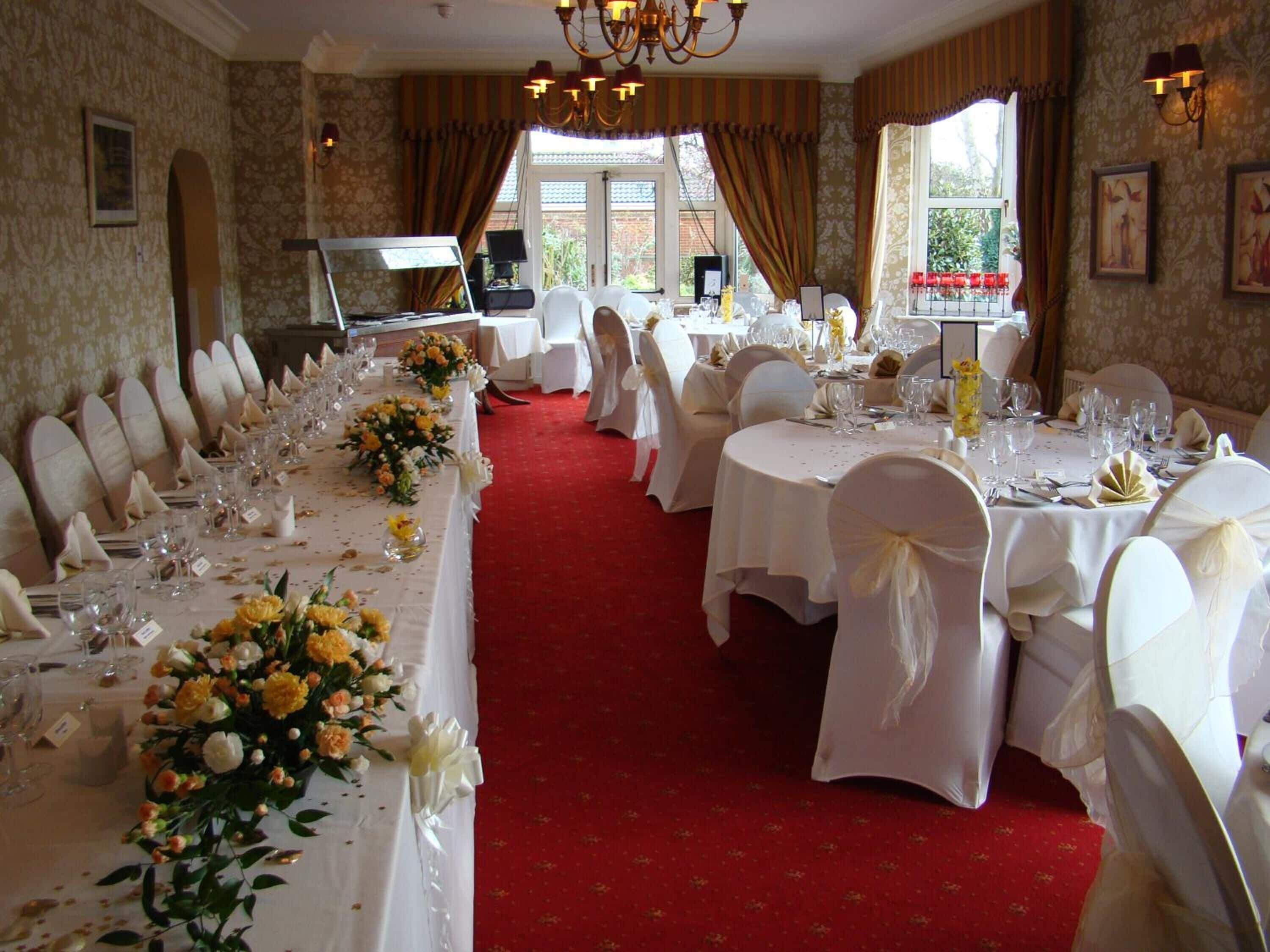 A banquet hall with long tables set for a formal event, featuring white tablecloths, floral centerpieces, and white chairs.