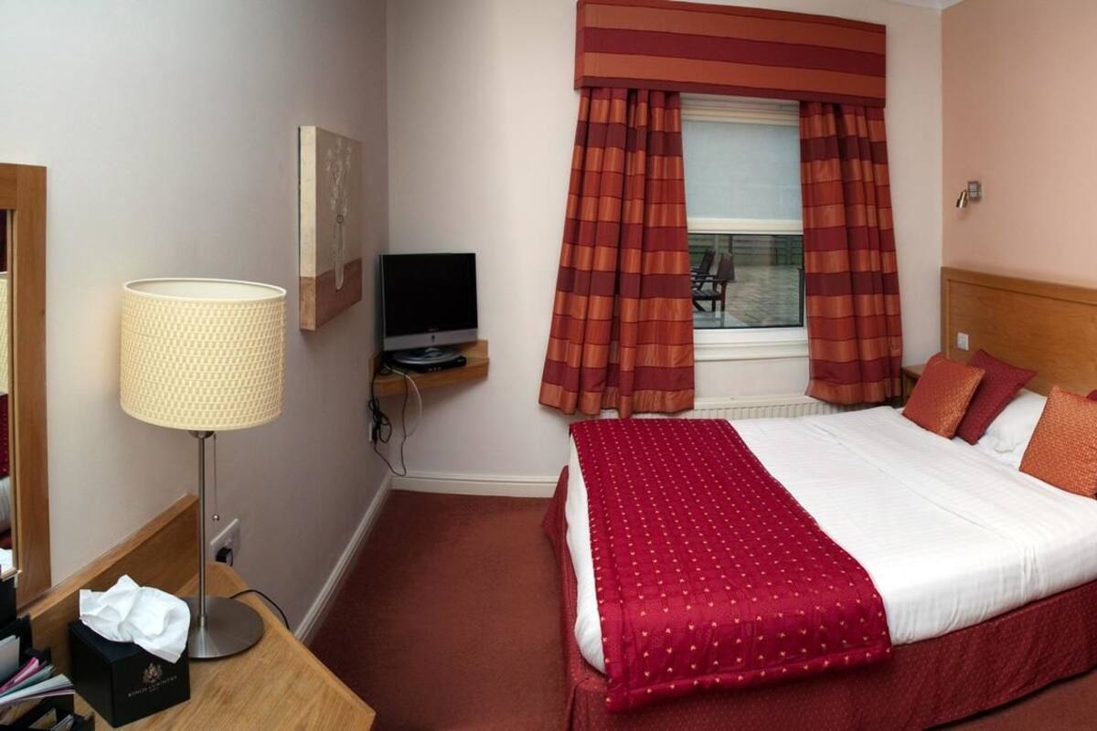 A hotel room with a bed, a desk with a lamp, a television, and a window with curtains.