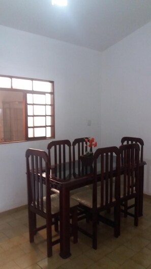 A dining area with a glass table surrounded by wooden chairs, a window with blinds, and a potted plant on the table. - Casa Mineira (Capitólio)