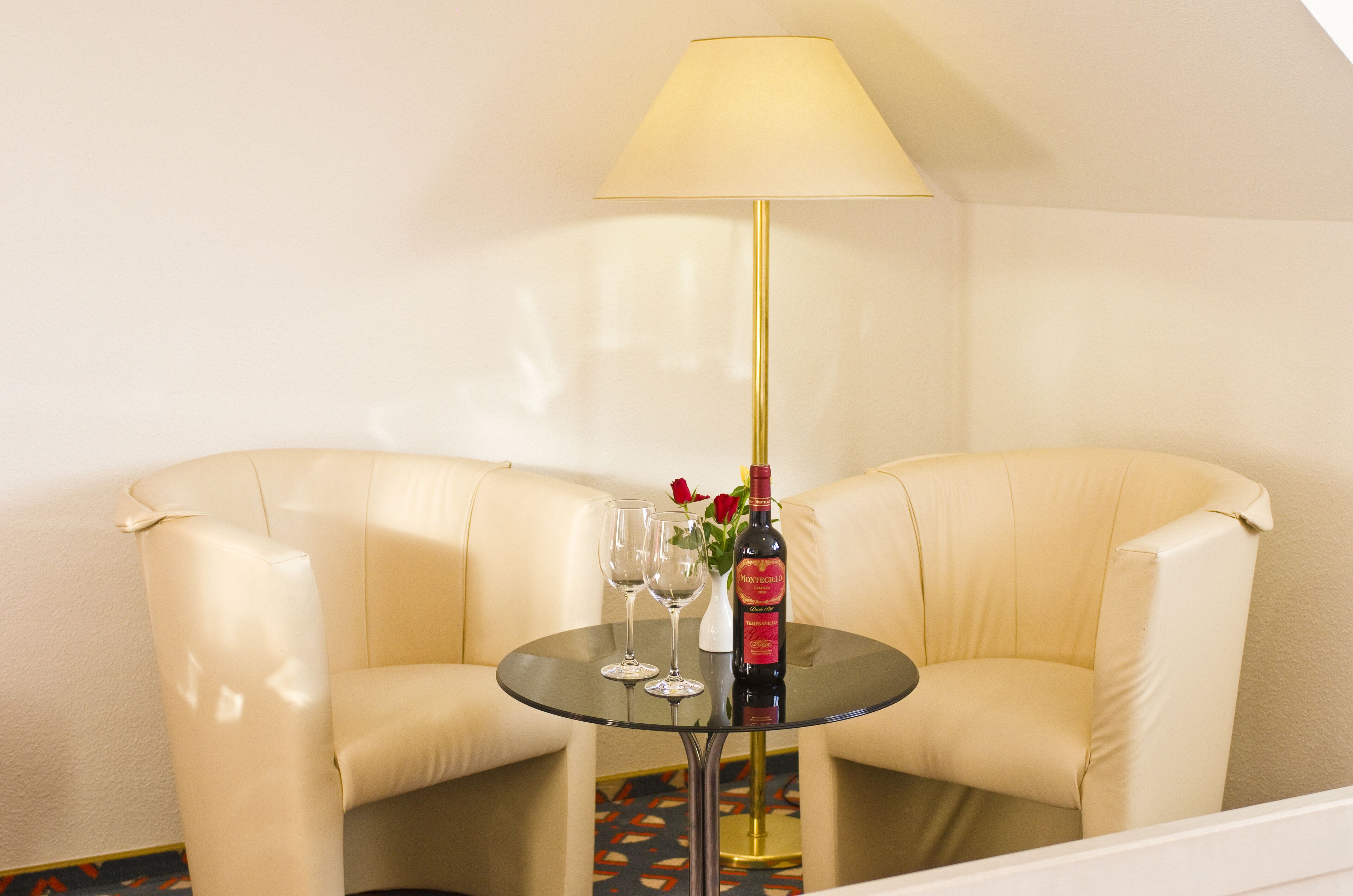 Une pièce avec une table ronde en verre, deux canapés en cuir blanc, une lampe, deux verres à vin et une bouteille de vin.