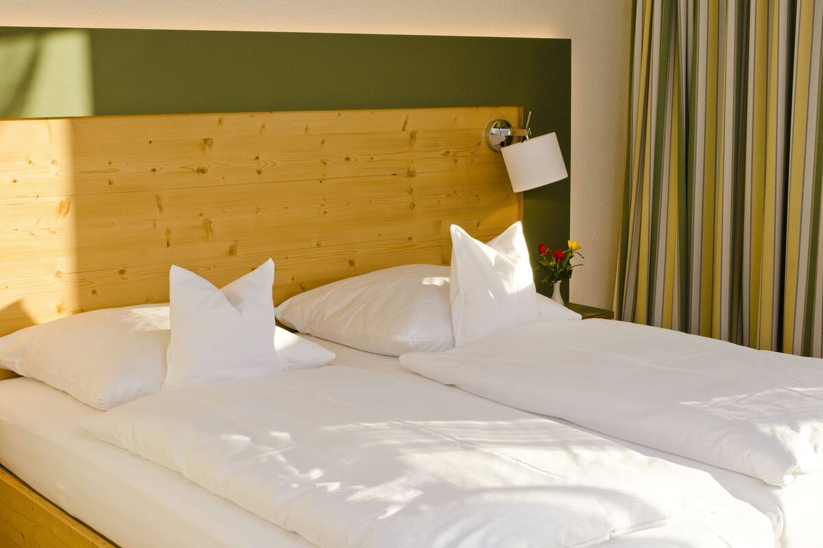 A double bed with white linens and pillows, a wooden headboard, a bedside table with a lamp, and a striped curtain.