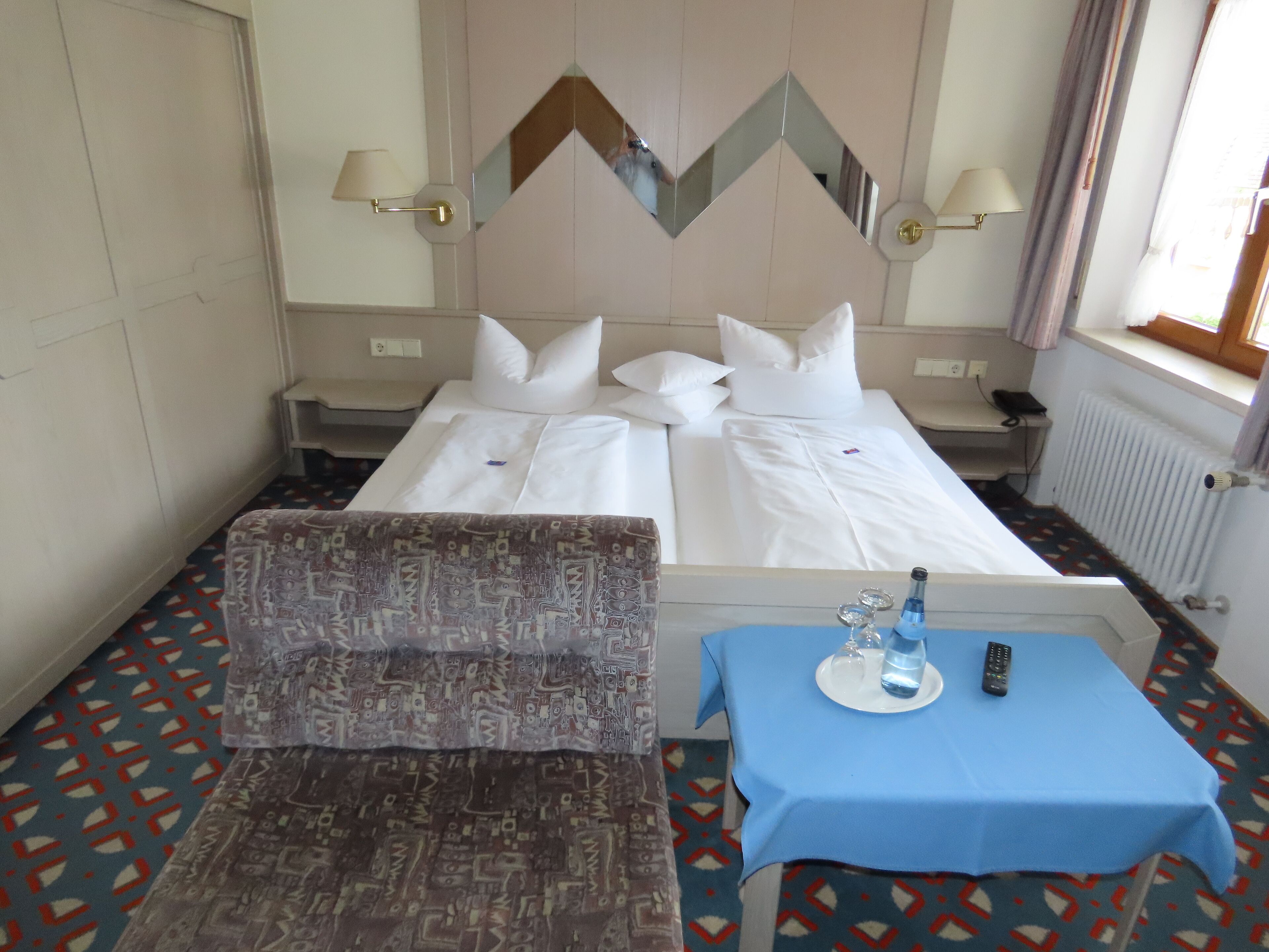 Une chambre d’hôtel avec un lit, une chaise, une table avec une bouteille et des verres, et un miroir au mur.