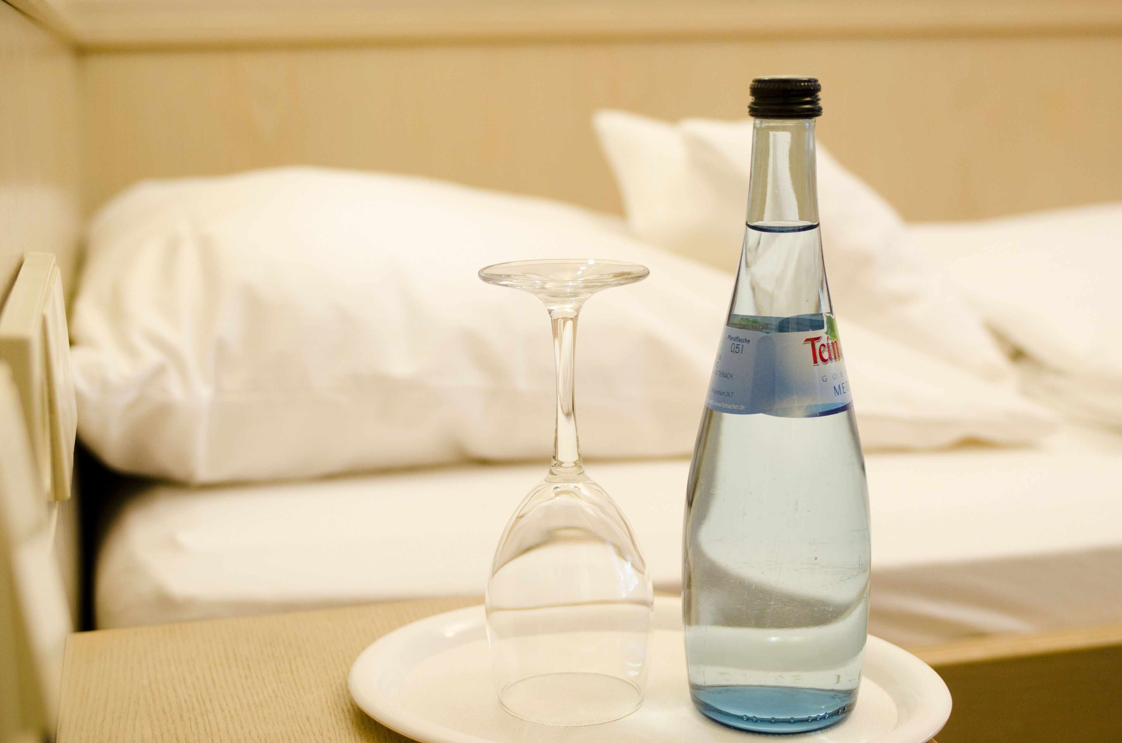 A bottle of water and an empty wine glass on a white plate on a bedside table in a hotel room.