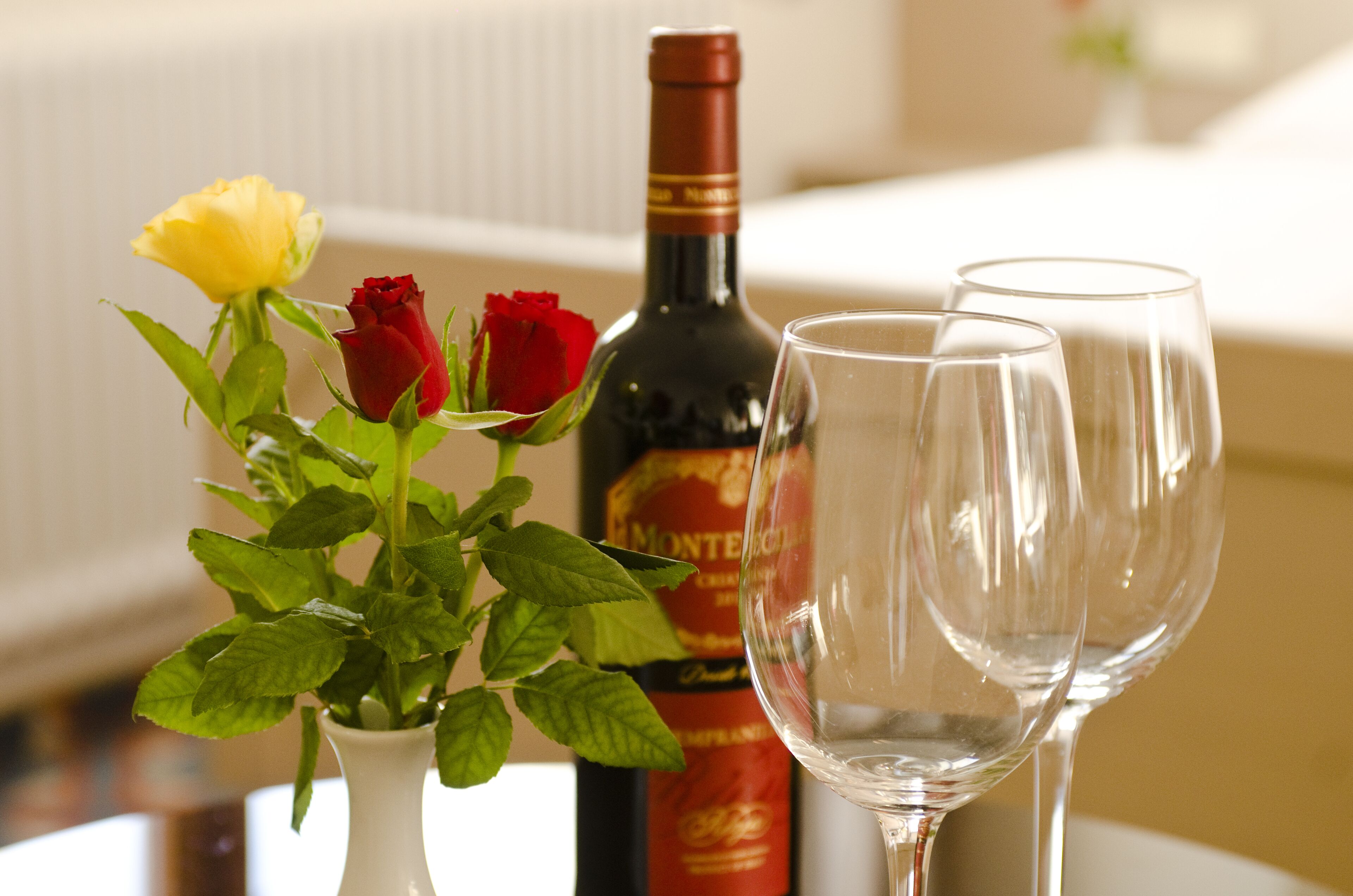 A table with a white vase containing a yellow rose and a red rose, a bottle of wine, and two empty wine glasses.