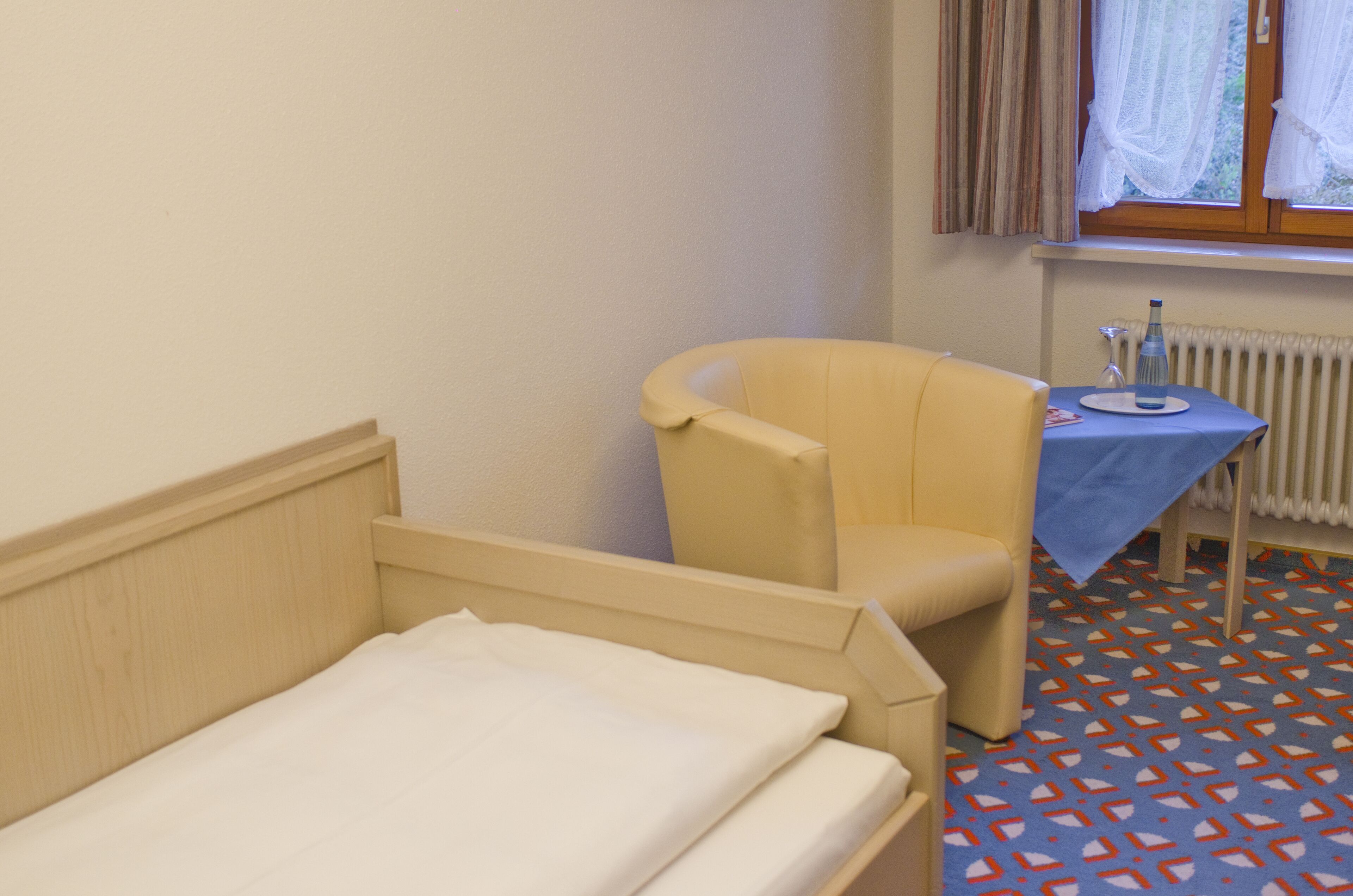 Une chambre d’hôtel avec un lit, une chaise, une petite table sur laquelle se trouve une bouteille, et une fenêtre avec des rideaux.