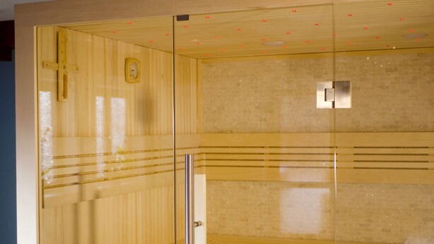 A sauna room with wooden paneling, a glass door, and a tiled wall. - Hotel Mutzel (Schluchsee)