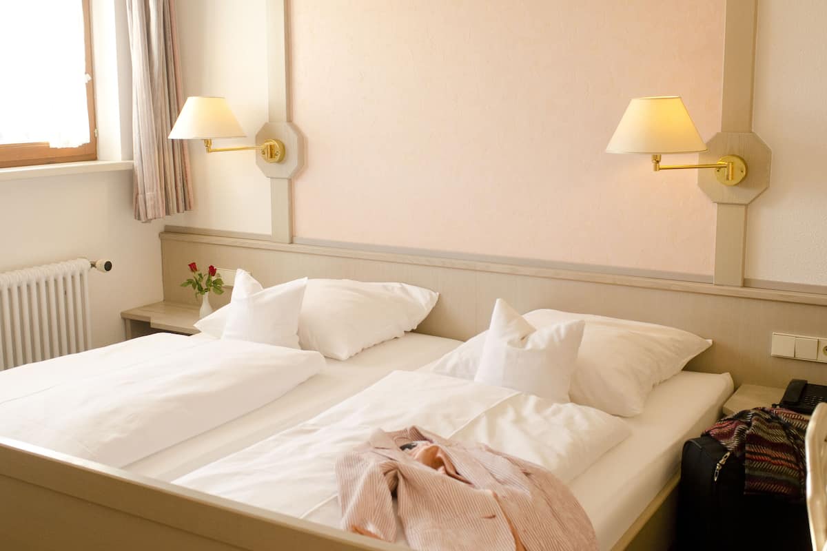 A hotel room with a bed, two wall-mounted lamps, a nightstand, a radiator, and a window with curtains.