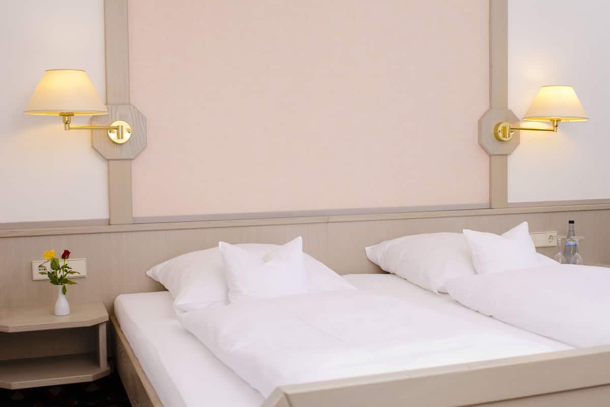 A hotel room with two beds, a nightstand with a vase of flowers, and wall-mounted lamps.