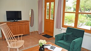 Una habitación con un armario de madera, televisión, una silla verde, una mesa de madera con un jarrón y una revista, y una ventana con cortinas.