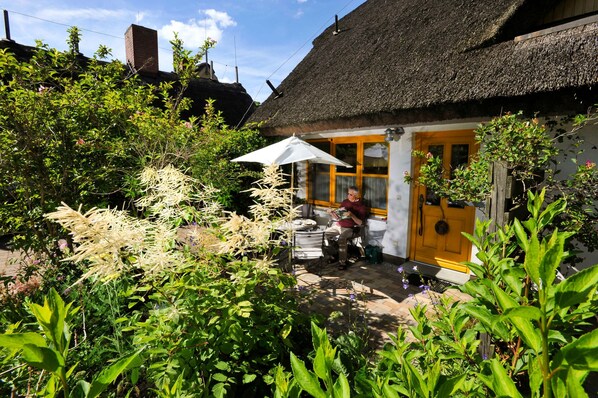 A thatched-roof cottage with a wooden door and a small patio area with a table and chairs.
