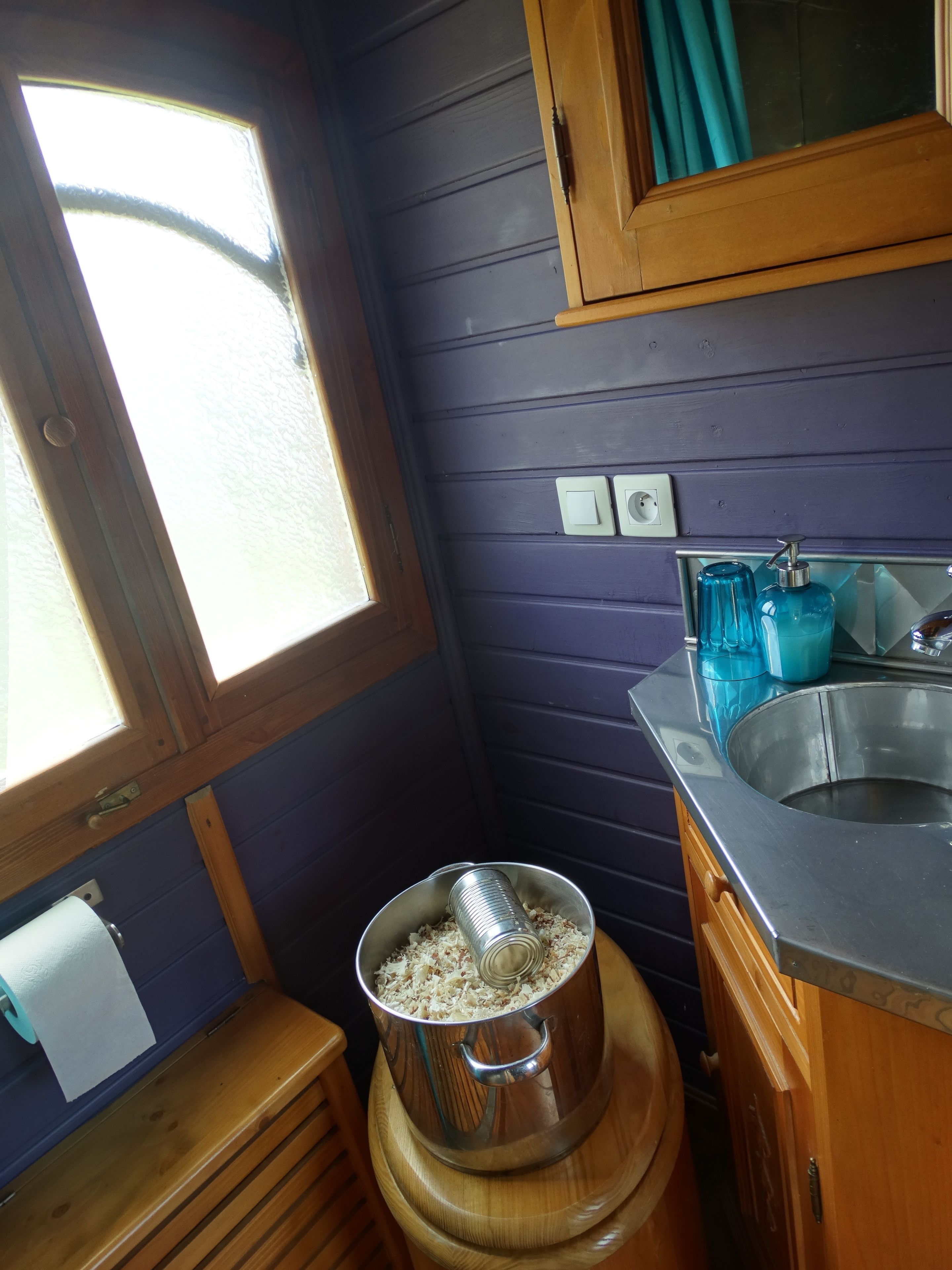 Cocina compacta con ventana, armarios de madera, un lavamanos de acero inoxidable y una olla sobre un taburete de madera.